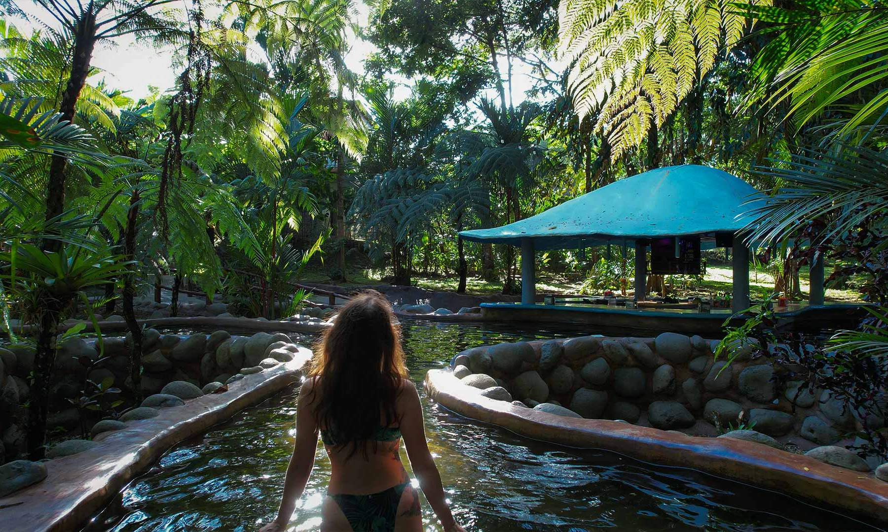Relaxing scene of a person enjoying the warm thermal waters amidst vibrant green foliage and sunlight at a natural hot spring resort